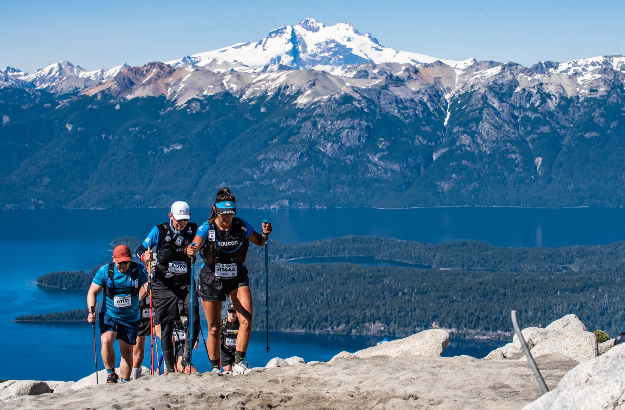 Three runners climb with poles, with the lake and the snowy range behind them