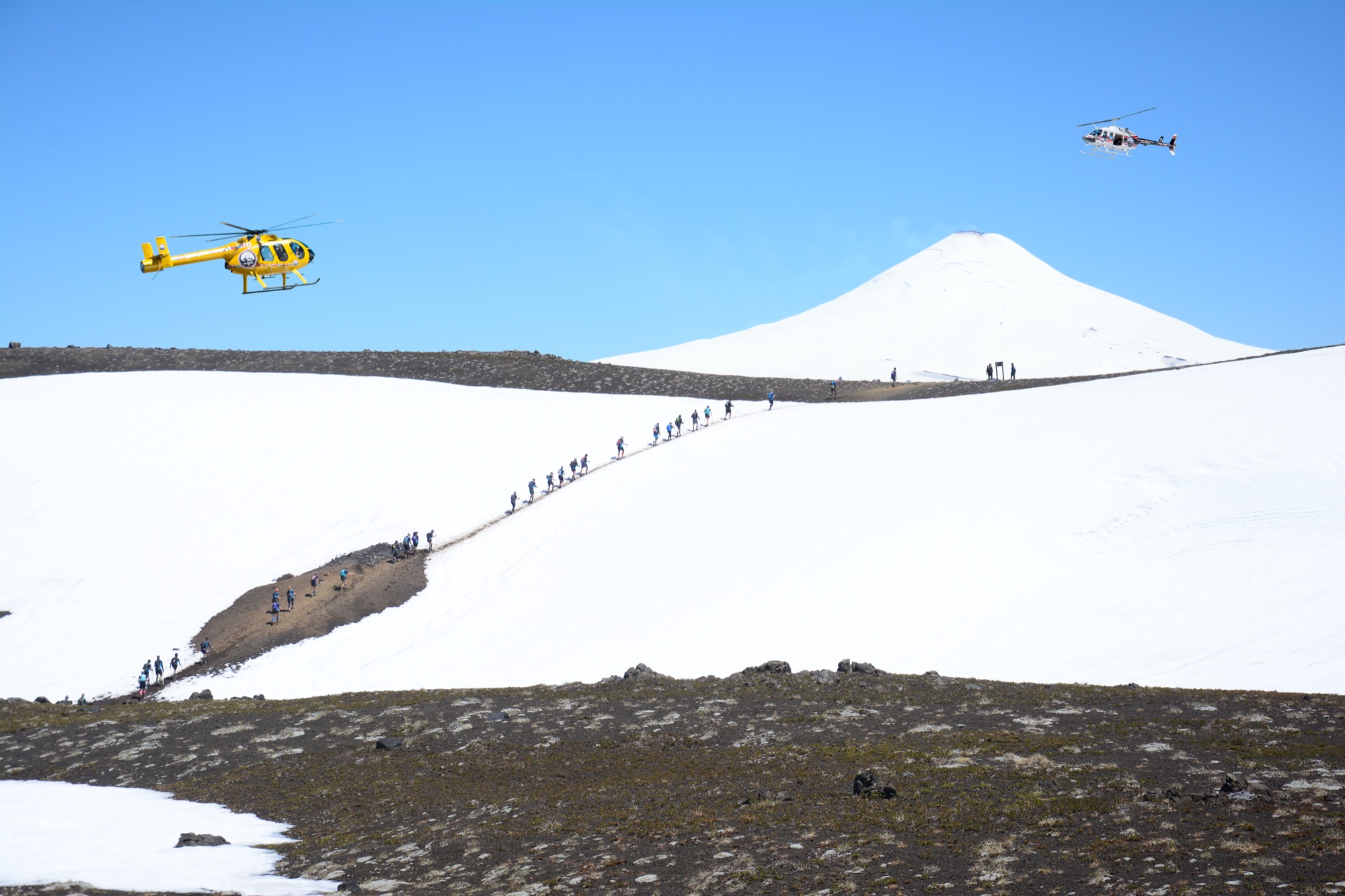 Two rescue helicopters fly over a line of runners crossing a snowfield toward a volcano