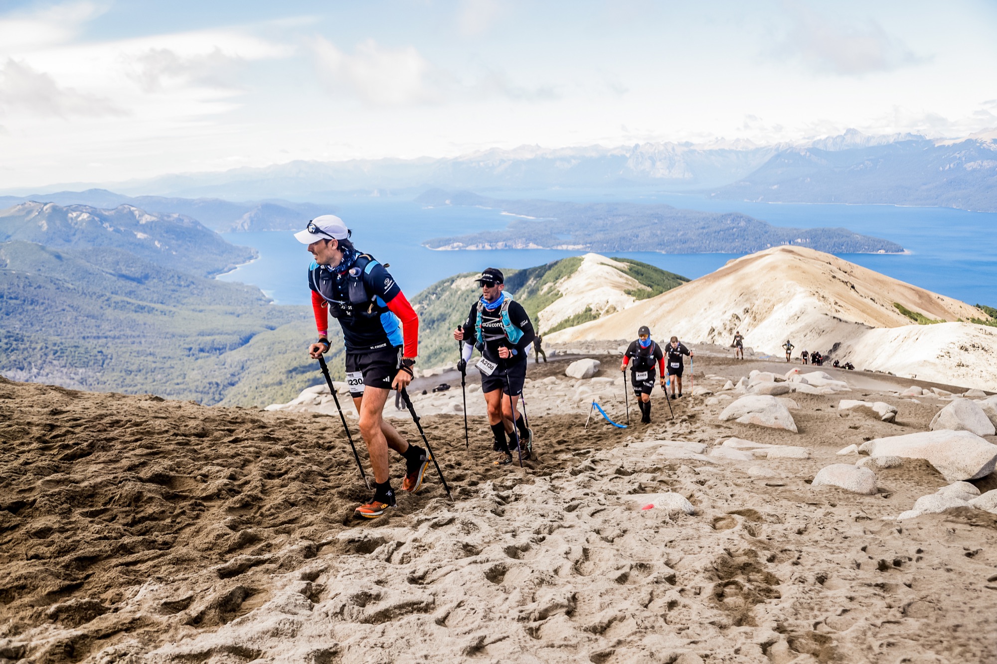 Runners climb a slope of volcanic sand with the lake in the background