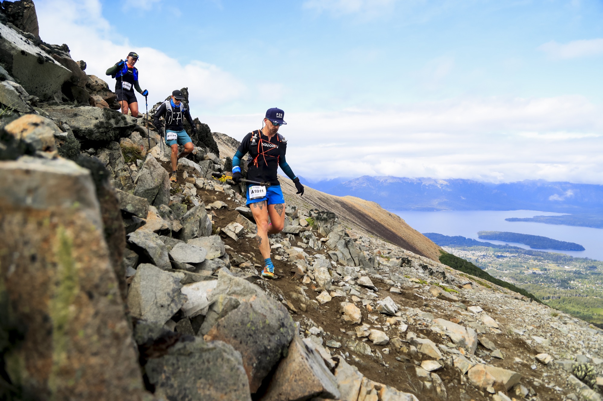 Runners scramble up a rocky slope with the lake in the background