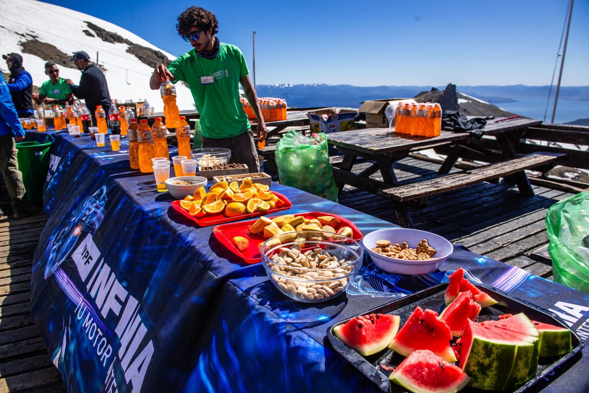 A volunteer prepares drinks at an aid station with fruit, crackers and drinks, facing the mountains and the lake